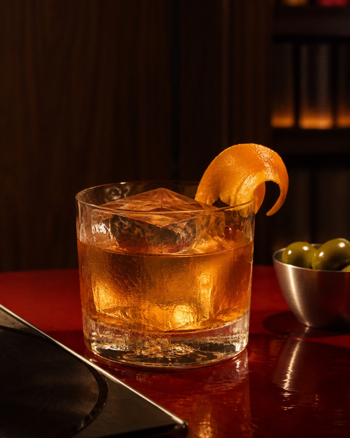An amber cocktail with clear ice and an orange peel on an oxblood lacquer table