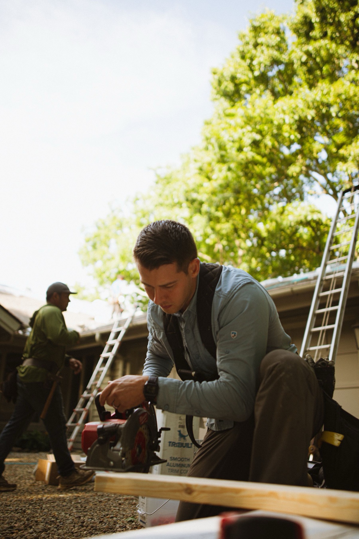 Two Ridgeline crew members at the truck on measure day — one cutting ridge vent at the sawhorse, one carrying a ladder to the house